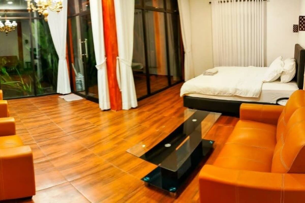 A modern hotel room with a wooden floor, orange sofas, a bed with white linens, and a large abstract painting on the wall.