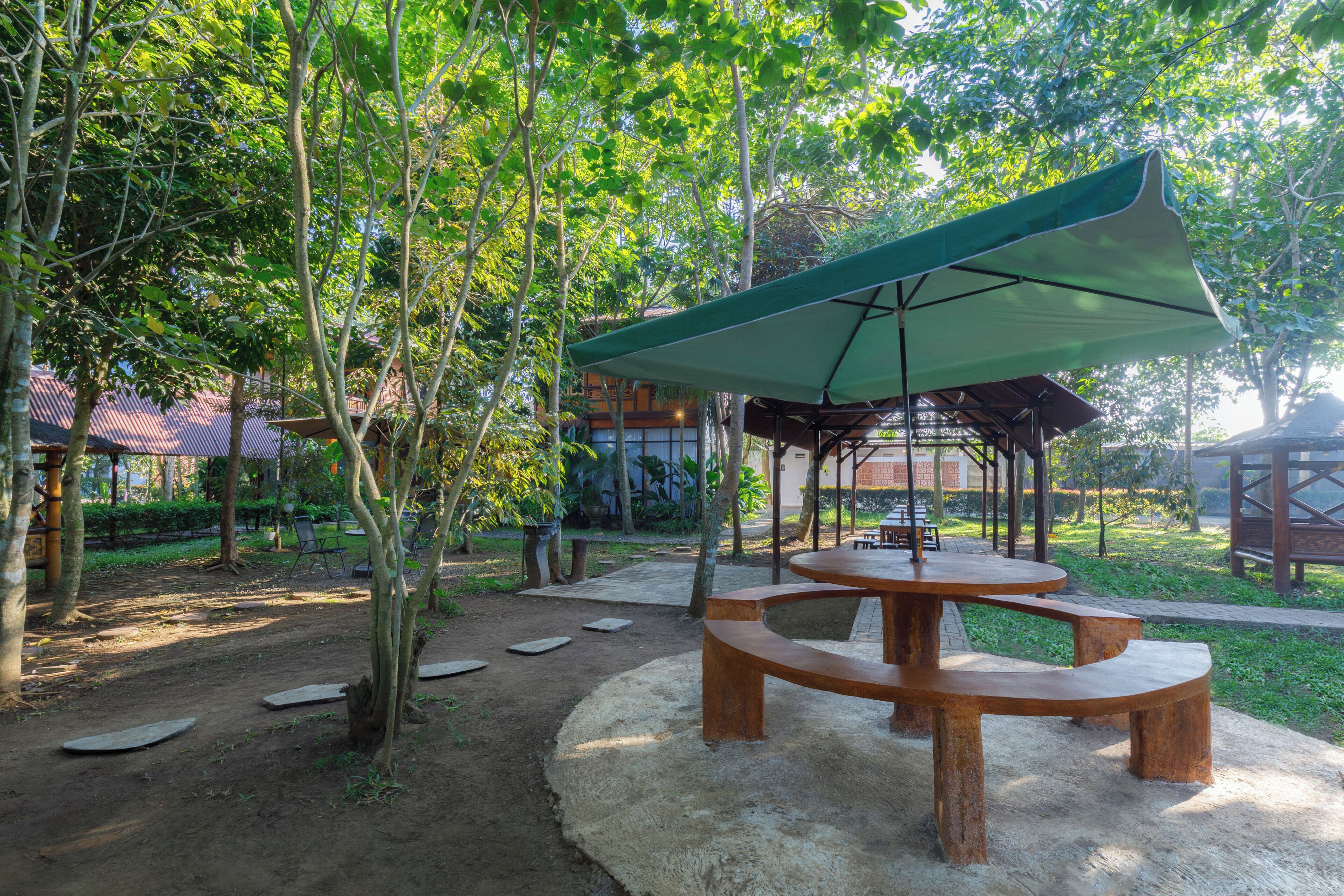 Ein Außenbereich mit Sitzgelegenheiten, darunter ein Holztisch und Bänke, umgeben von Bäumen und einem überdachten Pavillon.