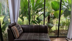 A dark brown leather sofa with white stitching and a striped pillow, positioned in front of a large window with a view of lush green plants and trees. - Ratu Roso Resort (Ulesari)