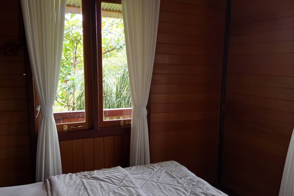 A wooden room with a bed, white curtains, and a window with a view of greenery.