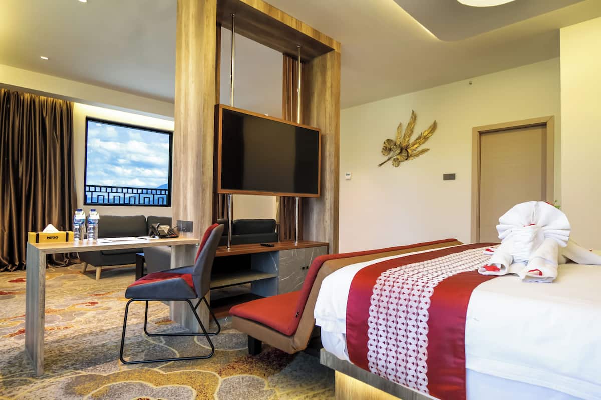 Ett hotellrum med en säng, ett skrivbord med en stol, en tv och ett stort fönster med gardiner.