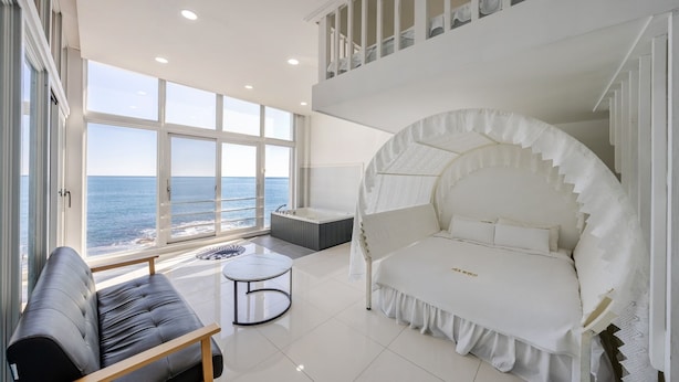 Un dormitorio moderno con un ventanal grande con vista al mar, una cama con dosel blanco, un sofá y una pequeña mesa redonda.