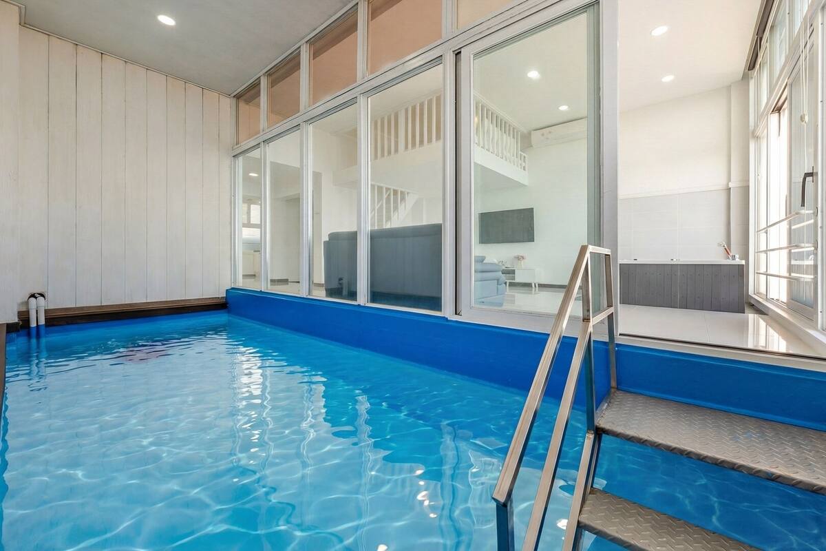 An indoor swimming pool with clear blue water, a ladder, and a view of a modern interior with white walls and large windows.