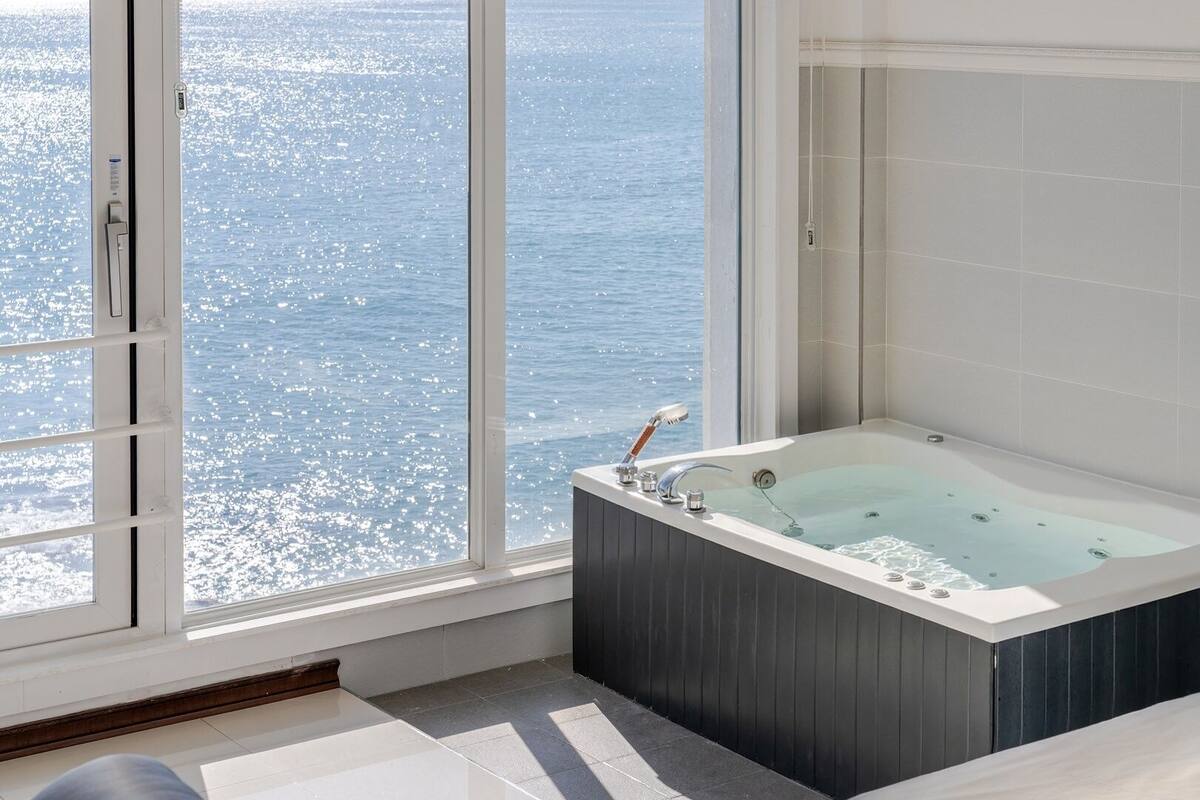 A modern bathroom with a large bathtub, a window overlooking the sea, and a shower area.