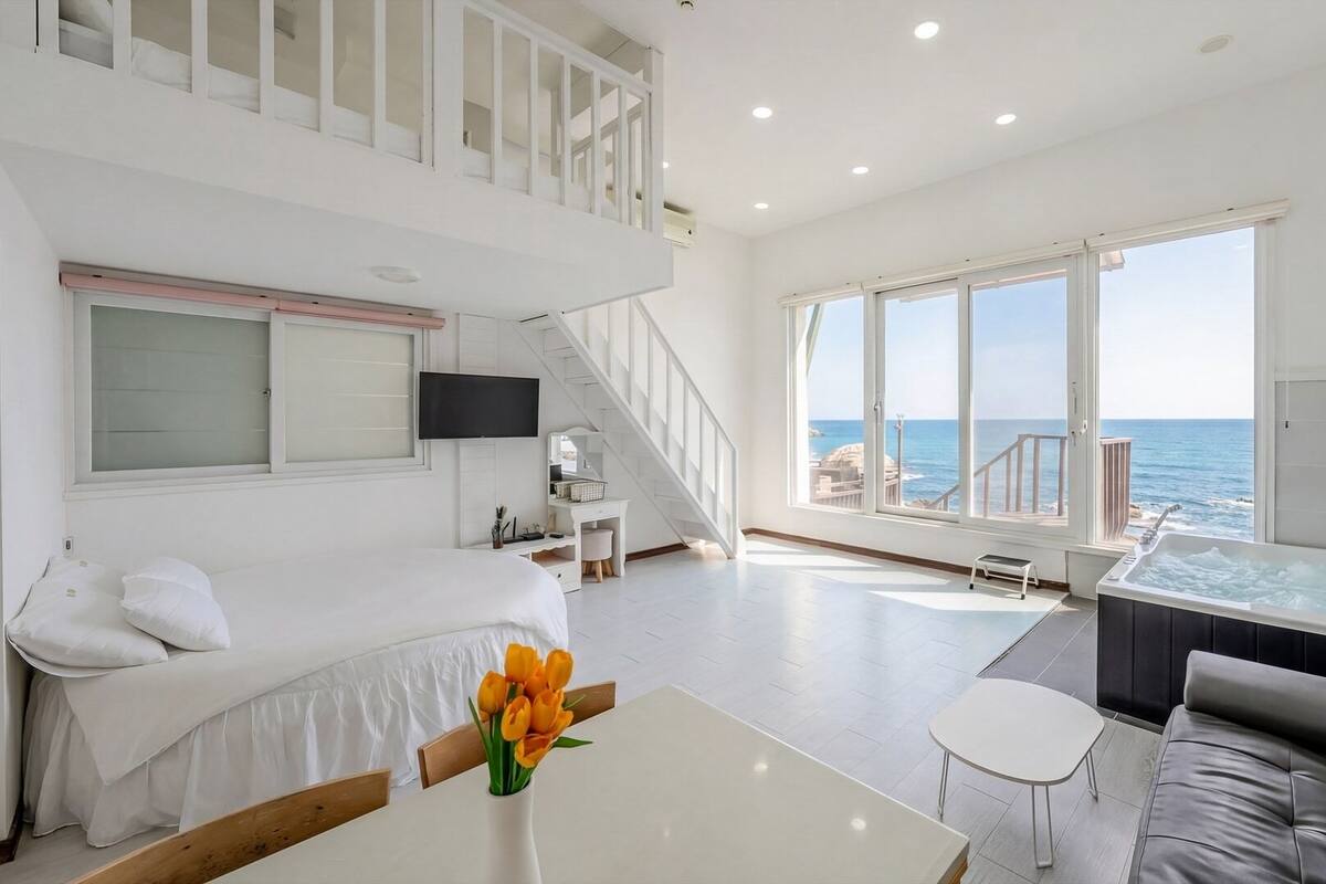 A modern hotel room with a balcony, a bed, a sofa, a table, and a jacuzzi.