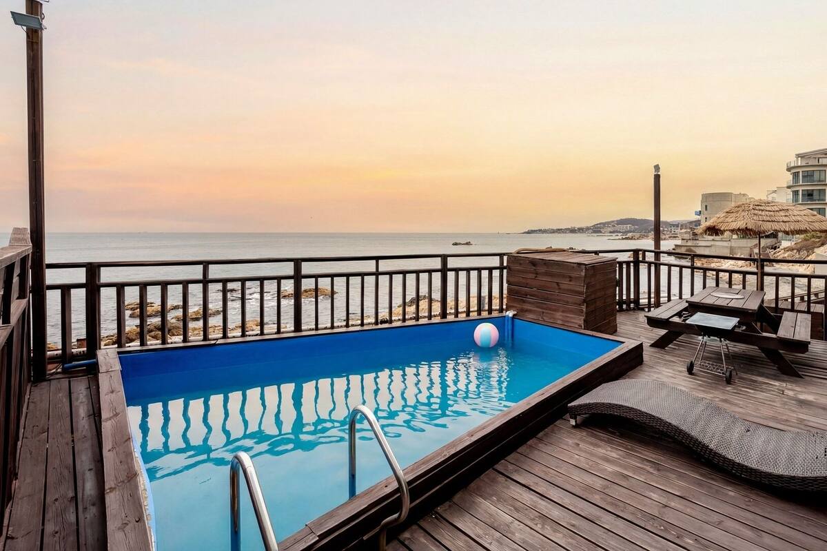A wooden deck with a swimming pool, a beach view, and a thatched-roof structure.