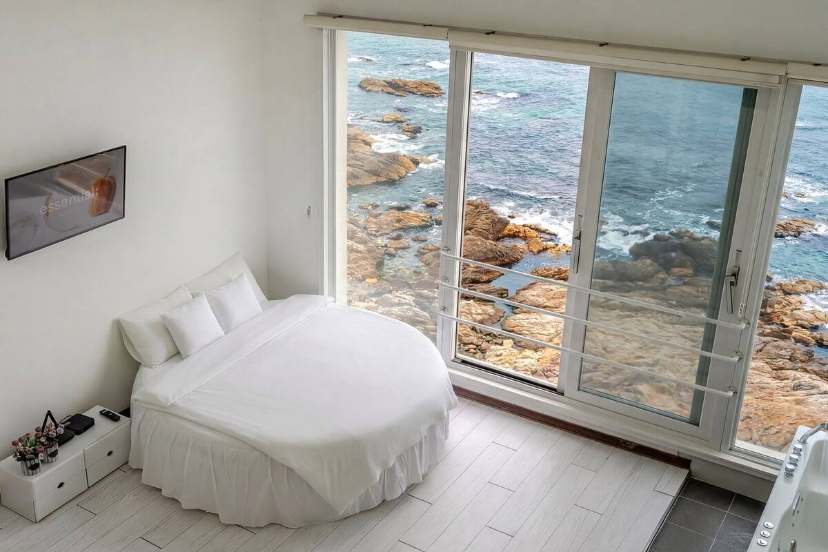 A bedroom with a bed, a bedside table, a nightstand, a framed picture, and a large window with a view of rocky coastline.