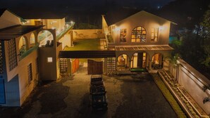 A well-lit courtyard with a covered seating area, a vehicle, and a building with multiple arches and a staircase. - The BaghVilla Pench FicusrootsExperience (Kurai)