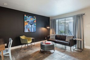 A modern living room with a black wall, a large window with curtains, a black sofa, a yellow chair, a round coffee table, and a painting. - 1BR The Montrose Hub, Downtown, Med Center 11 (Houston)