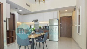 A modern kitchen with white cabinets, a stainless steel refrigerator, and a dining area with a wooden table and chairs. - Bloomfields Modern 1Bedroom In Wavez (Dubai)
