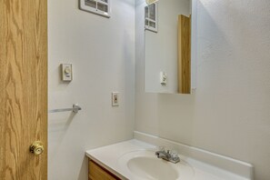 Un baño con puerta de madera, paredes blancas, un lavamanos blanco y un espejo.