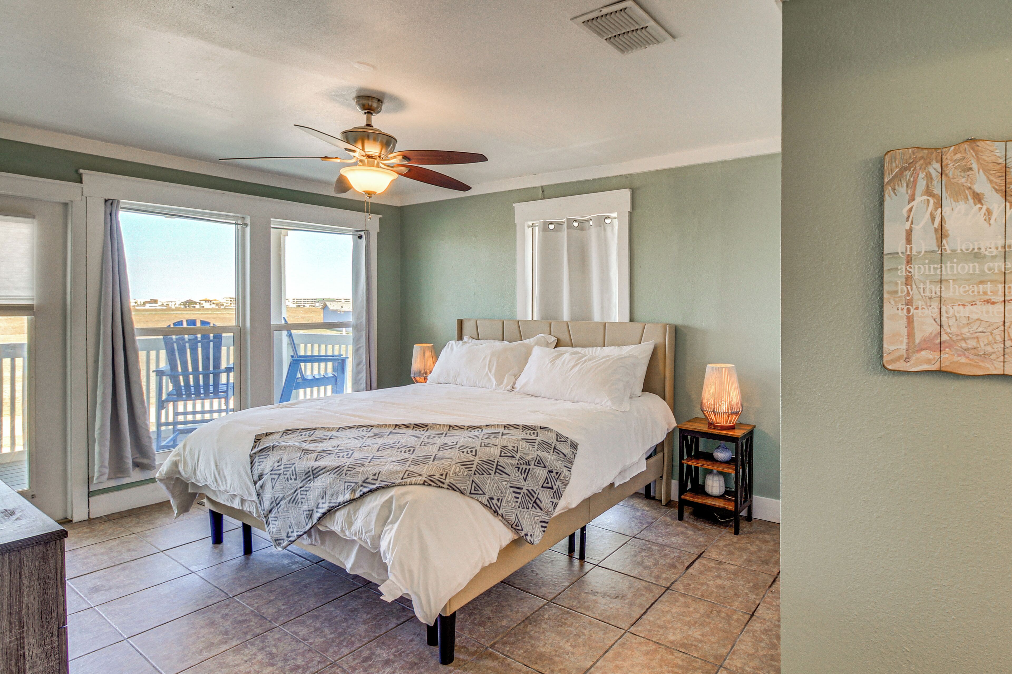A bedroom with a bed, a ceiling fan, a nightstand with a lamp, and a view of the outside.