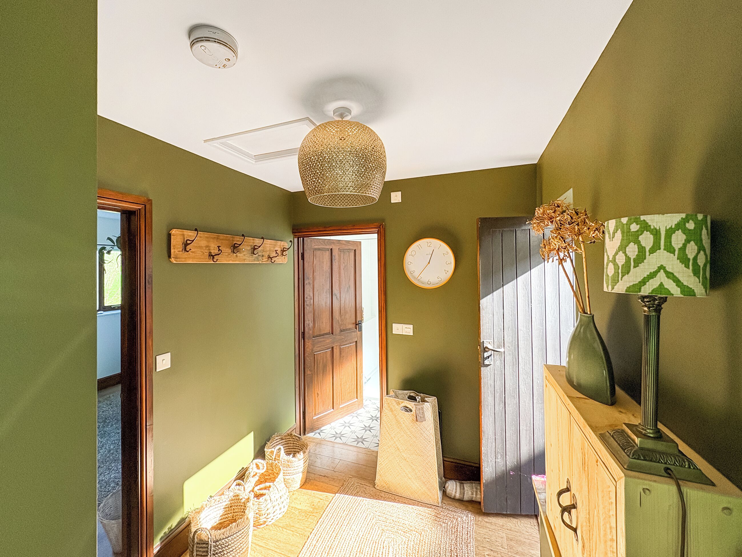 A hallway with a wooden door, a wall clock, a hanging light fixture, and a lamp on a cabinet.