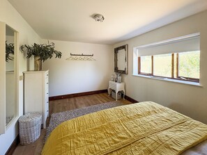 Ein Schlafzimmer mit Bett, Kommode, Spiegel, Pflanze und Fenster.