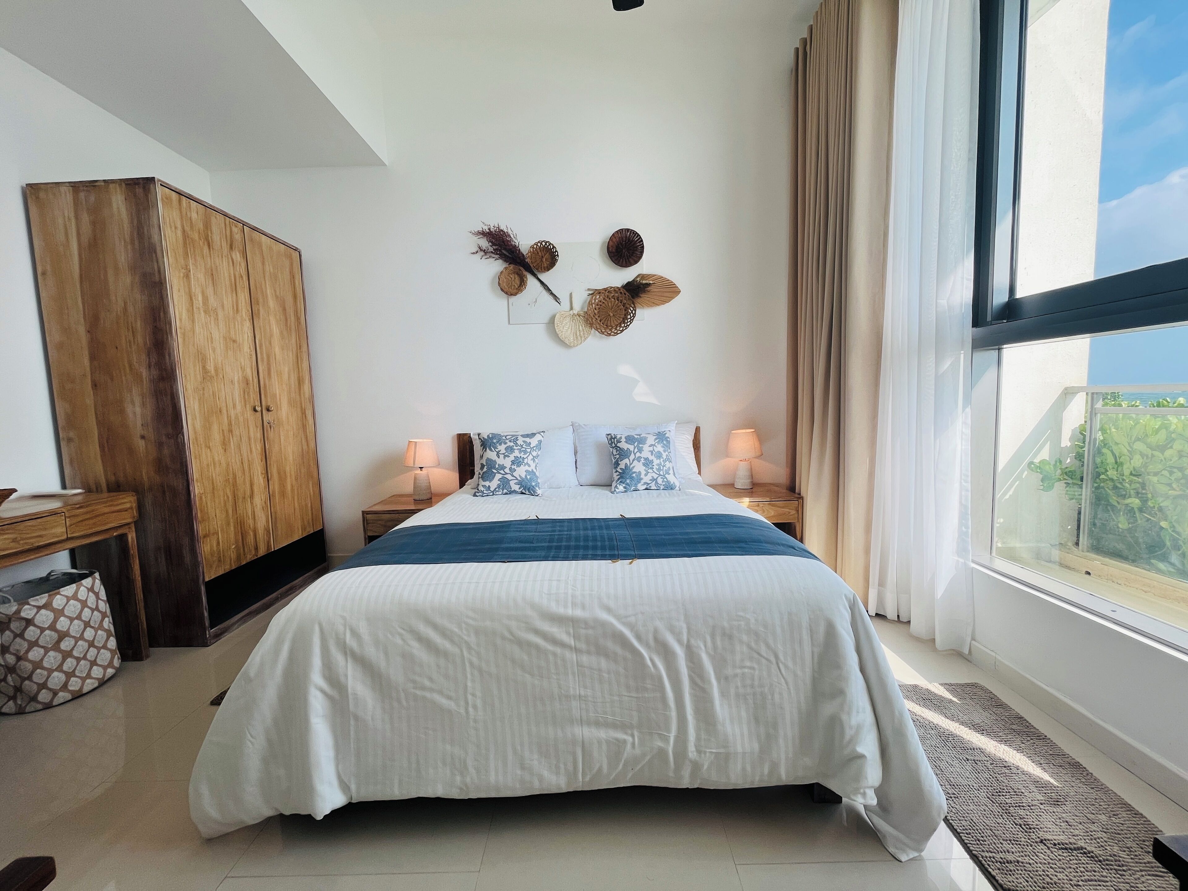 A bedroom with a large bed, wooden wardrobe, bedside lamps, and a view of greenery through the window.
