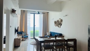 A modern living room with a dining area, a large window with curtains, a ceiling fan, and a wall-mounted decorative piece. - C-5 Oceanfront Condos  (Galle)