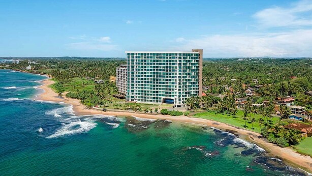 A high-rise hotel with a modern design, located near a beach with clear blue waters and lush greenery. - C-5 Oceanfront Condos  (Galle)