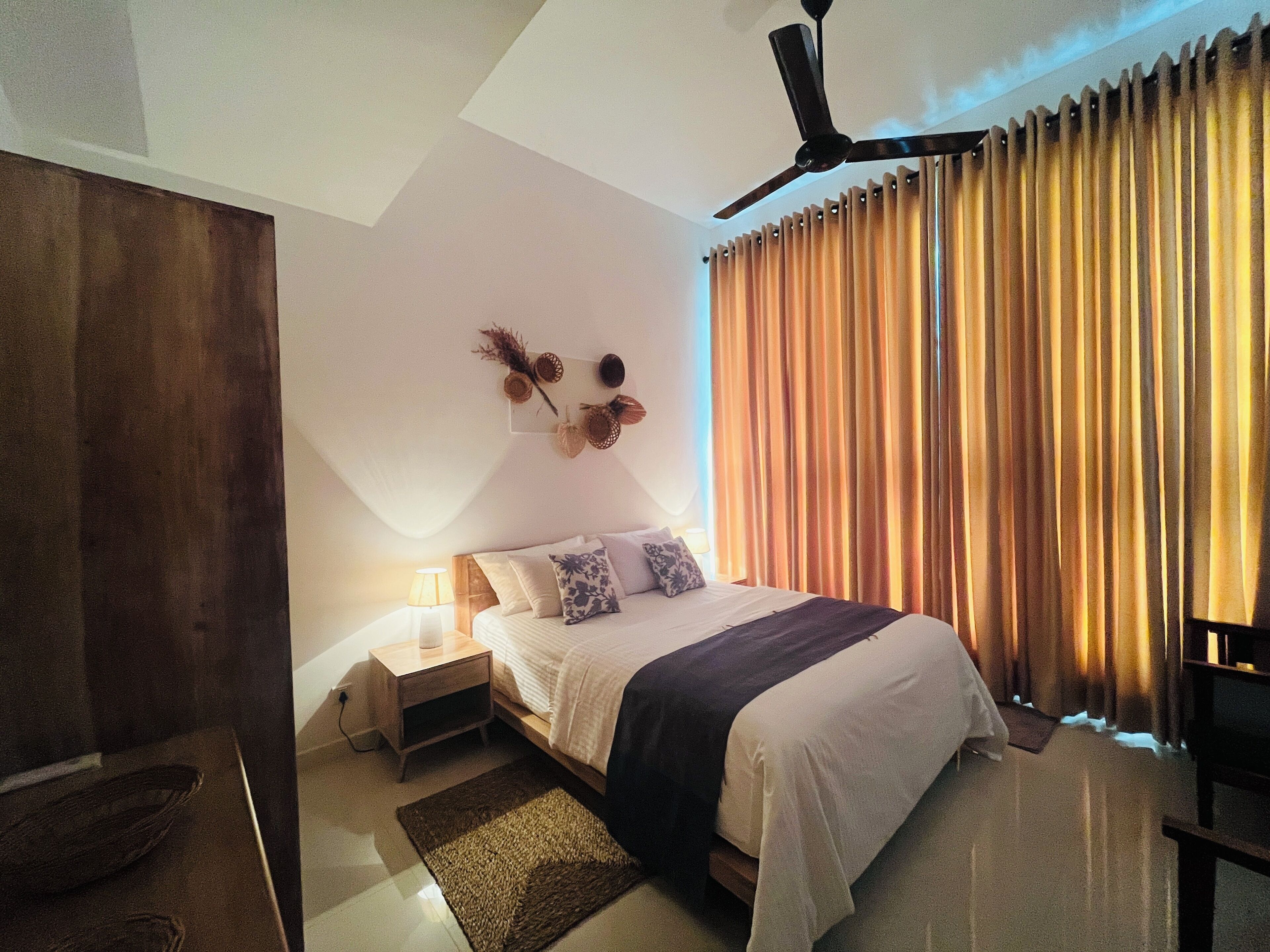 Un dormitorio con cama, mesitas de noche, ventilador de techo y cortinas.