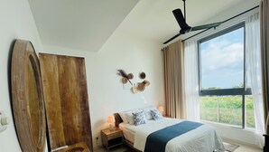 A bedroom with a bed, bedside lamps, a wooden nightstand, a large window with curtains, and a ceiling fan. - C-5 Oceanfront Condos  (Galle)