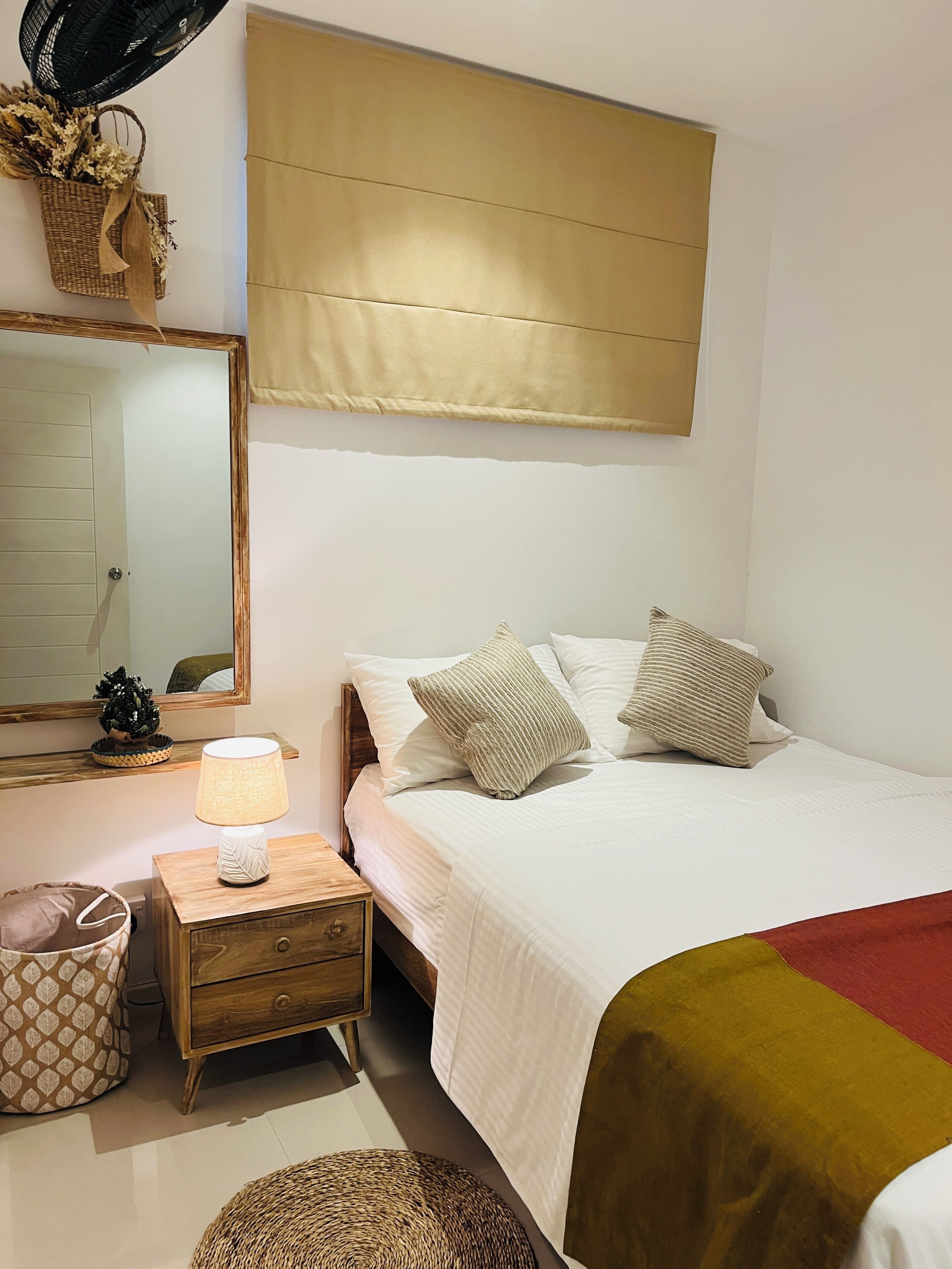 A bedroom with a bed, a bedside table with a lamp, a nightstand, a small potted plant, a mirror, and a wall-mounted fan.
