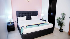 A neatly made bed with a black headboard, two bedside tables with lamps, a potted plant, and a closed door. - Fortune Home Service Apartment J-171 (New Delhi)