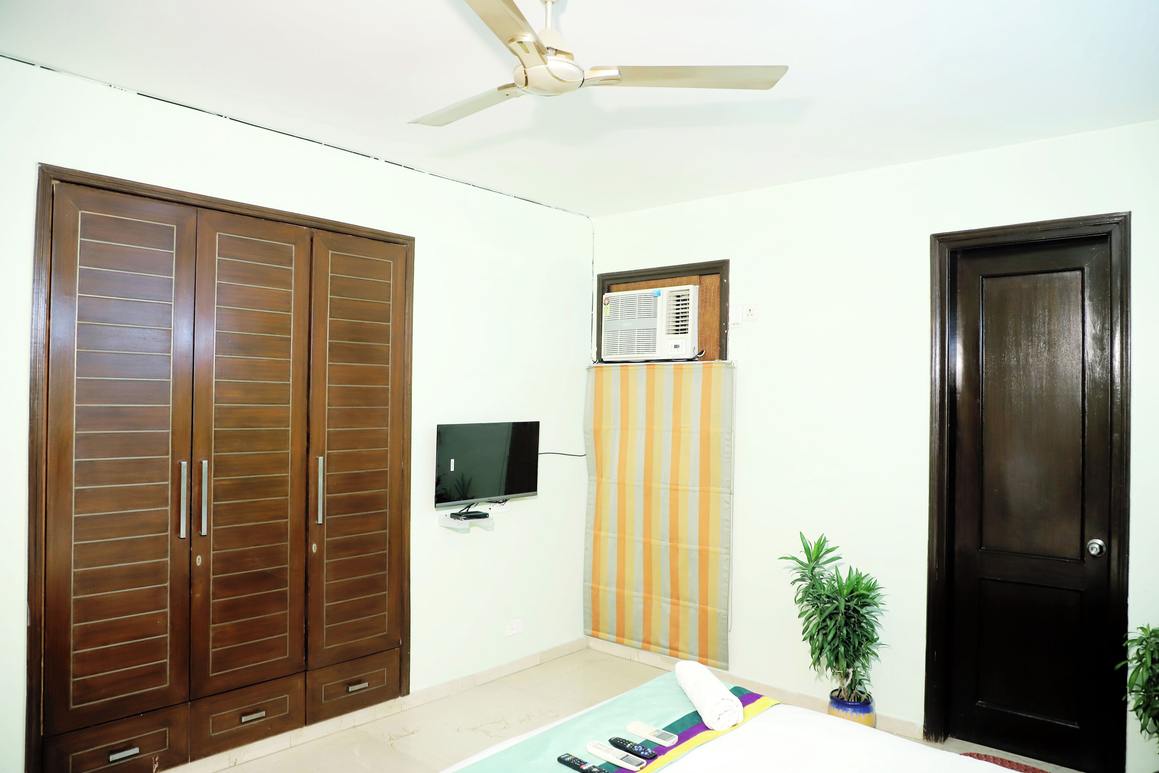 A bedroom with a ceiling fan, wardrobe, TV, air conditioner, striped curtain, and a potted plant.