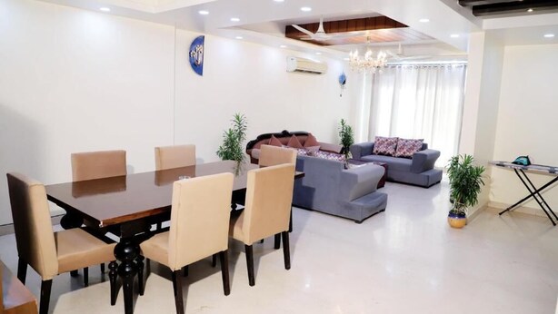 A modern living room with a dining area, featuring a wooden dining table, beige chairs, a grey sofa, and a blue armchair. - Fortune Home Service Apartment J-171 (New Delhi)
