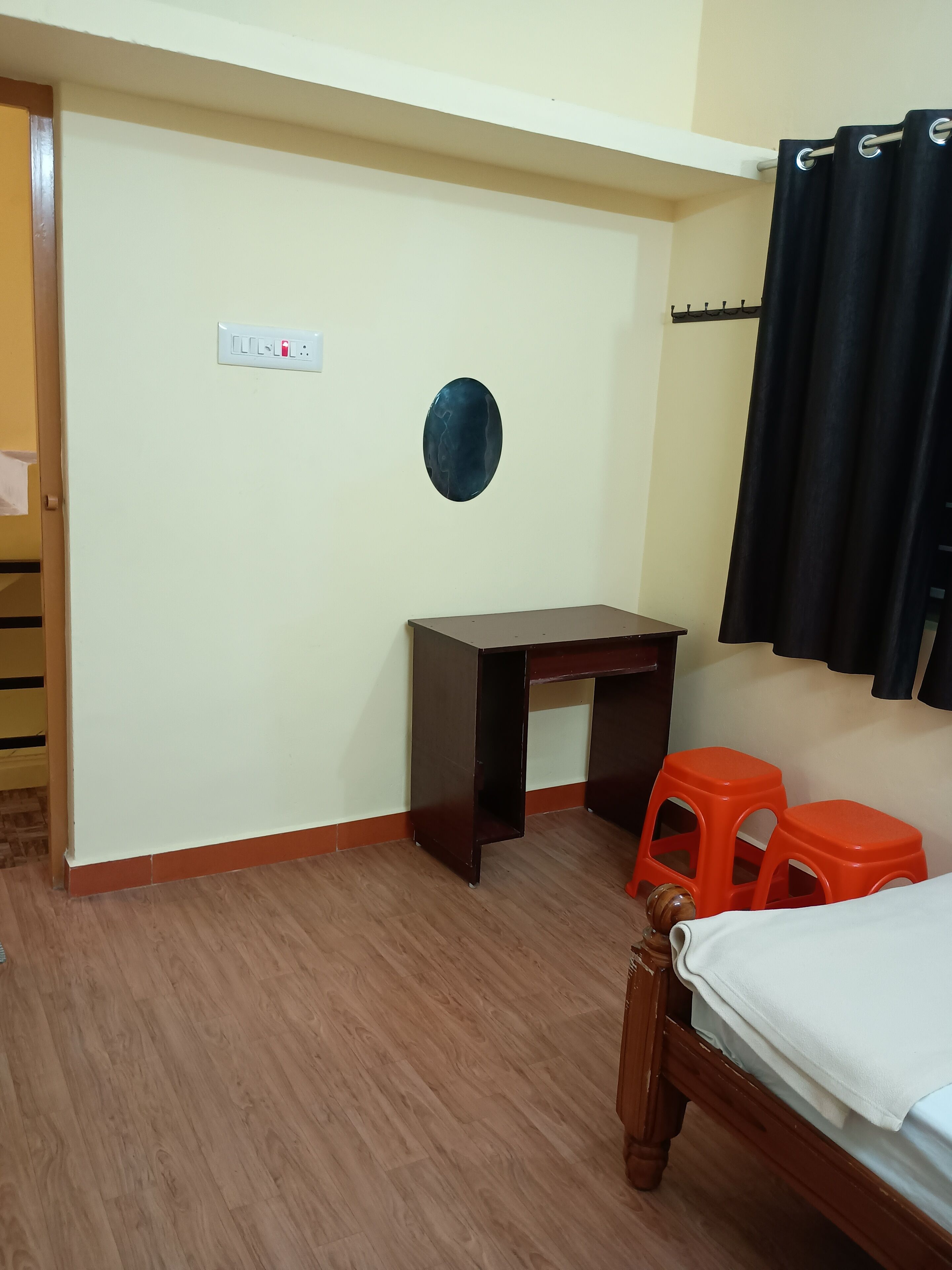 A room with a wooden desk, two orange stools, a bed with a white cover, a black curtain, and a wall-mounted light switch.