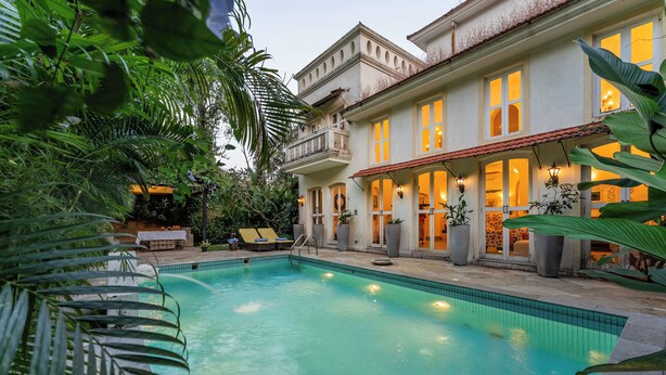 Piscina com espreguiçadeiras, cercada por vegetação exuberante e um edifício grande com várias janelas e varandas.