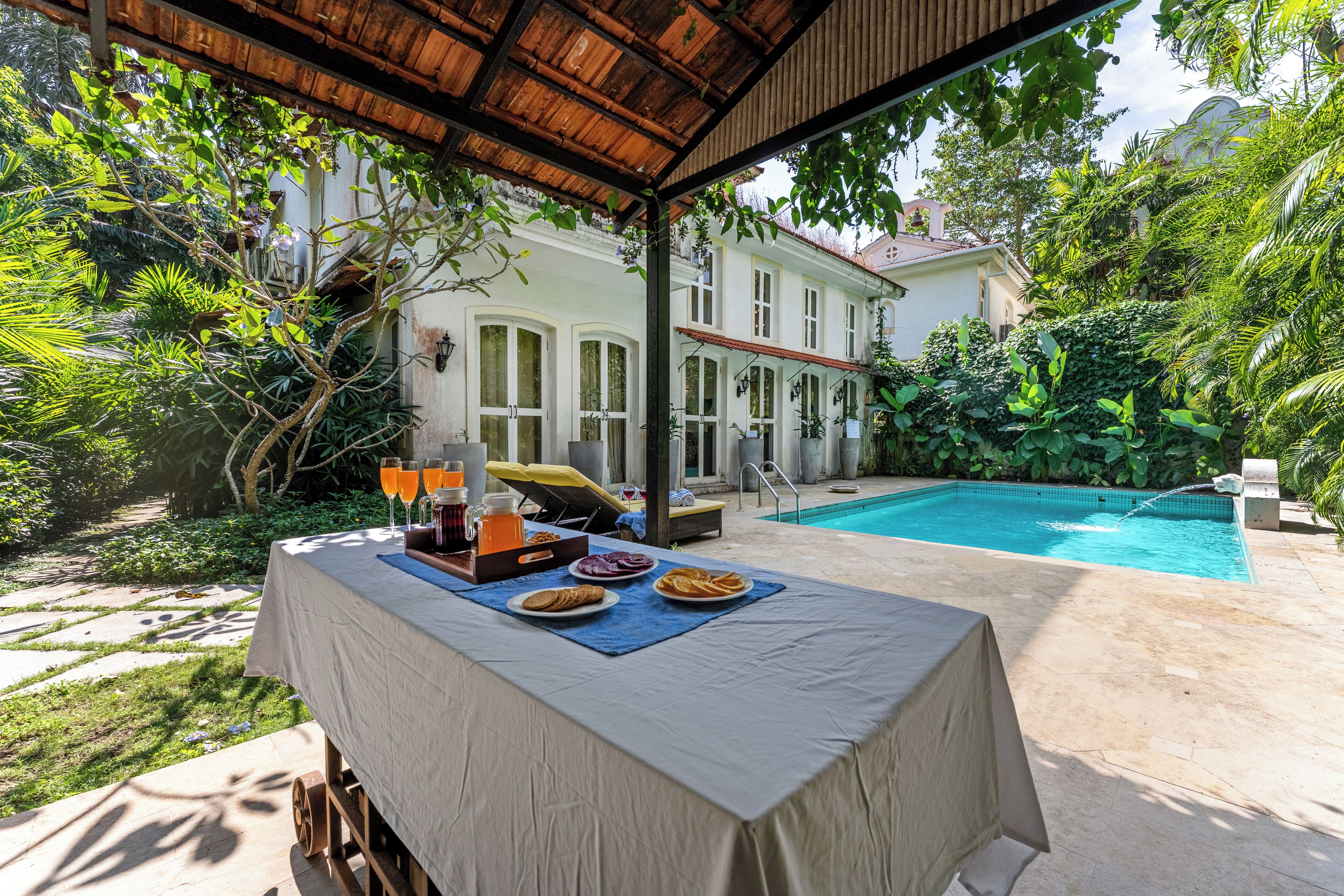 Uma área de refeições coberta ao ar livre, com uma mesa posta para o café da manhã, com vista para uma piscina e vegetação exuberante.