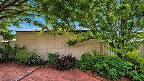 A brick wall with a garden area, including various plants and a paved walkway. - Sal’s Cottage - Charming Coastal Cottage in Busselton (West Busselton)