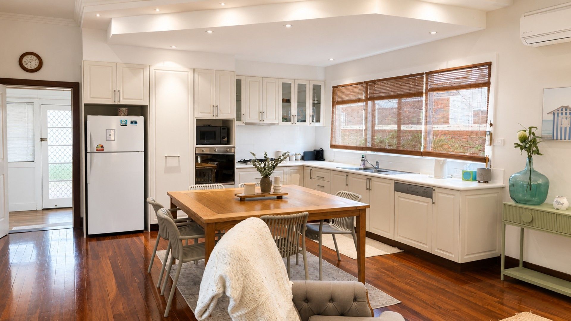 A modern kitchen with white cabinetry, a wooden dining table, and a window with blinds.