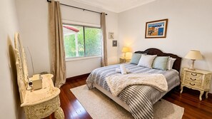 A bedroom with a bed, bedside tables, a mirror, a window with curtains, and a framed picture on the wall. - Sal’s Cottage - Charming Coastal Cottage in Busselton (West Busselton)