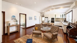 A spacious living area with a wooden floor, a dining table, two armchairs, and a kitchen in the background. - Sal’s Cottage - Charming Coastal Cottage in Busselton (West Busselton)