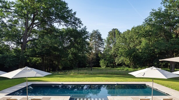 A swimming pool with a checkered floor, surrounded by lounge chairs and umbrellas, set against a backdrop of trees.