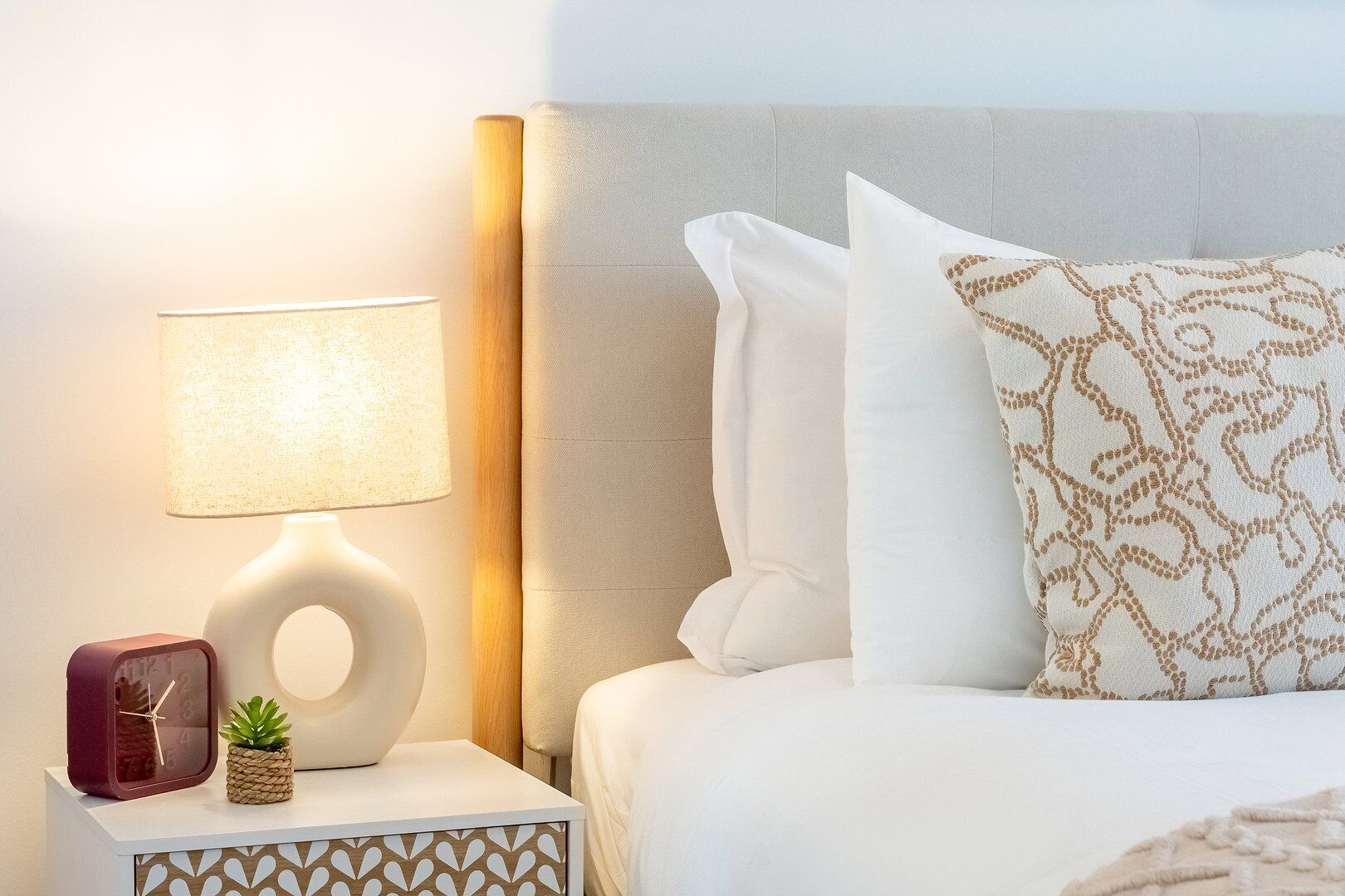 A neatly arranged hotel room with a bed, a bedside lamp, a clock, and a small potted plant.