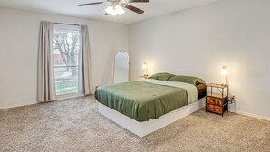 A bedroom with a bed, a ceiling fan, a nightstand with a lamp, and a window with curtains. - DFW Home Base! Quiet Suburb Stay w/ Yard (Carrollton)