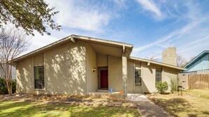 A single-story house with a red door, a covered porch, and a small front yard. - DFW Home Base! Quiet Suburb Stay w/ Yard (Carrollton)