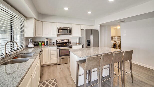 A modern kitchen with white cabinets, stainless steel appliances, a granite countertop, and a bar stool seating area. - DFW Home Base! Quiet Suburb Stay w/ Yard (Carrollton)