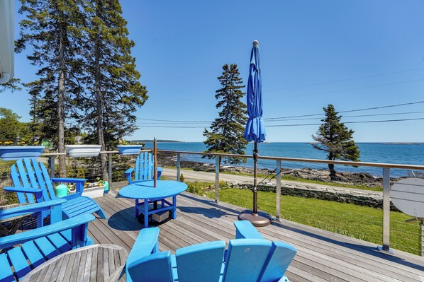 Een terras met blauwe Adirondack-stoelen en een tafel, met uitzicht op een rotsachtige kustlijn en de oceaan.