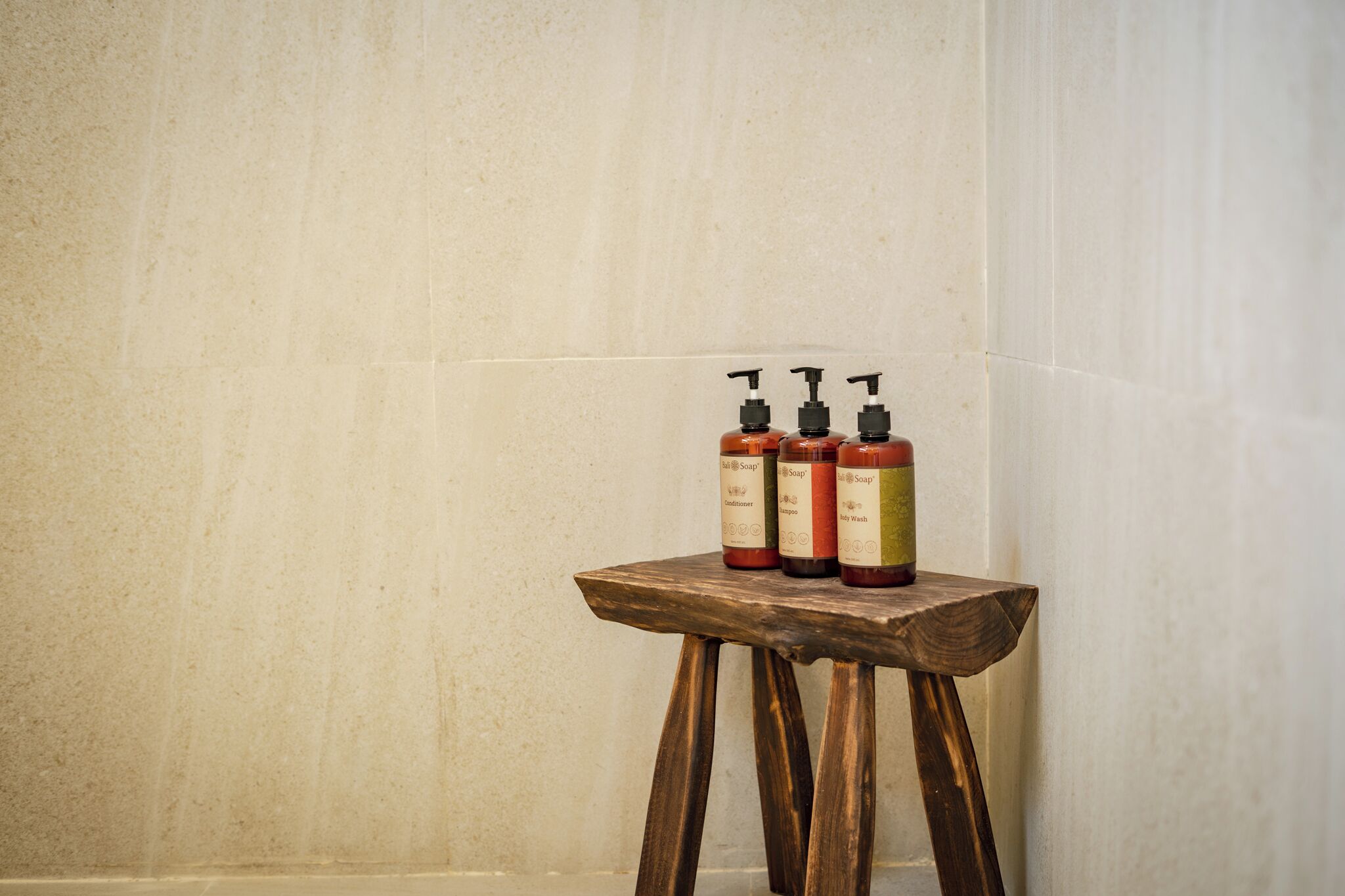 A wooden stool with three bottles of soap against a tiled wall.
