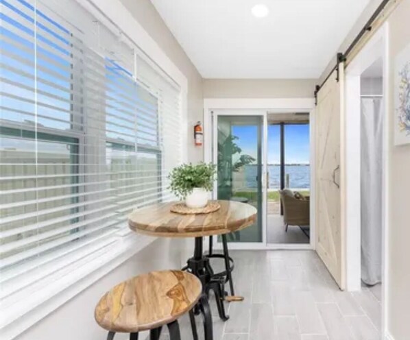 A narrow balcony with a wooden table and two stools, a potted plant, and a view of the sea through a glass door.