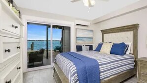 A bedroom with a bed, a ceiling fan, a view of the ocean through a sliding door, and a small balcony with a chair. - Bayfront Condo, Prime Beach Access, Manasota Key (Englewood)
