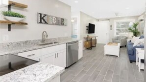 A modern kitchen with a built-in stove, white cabinetry, and a granite countertop. There is a dining area with a white table and chairs, and a living room with a sofa and a TV. - Bayfront Condo, Prime Beach Access, Manasota Key (Englewood)