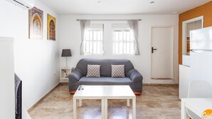 A living room with a grey sofa, a white coffee table, and a TV. There are two windows with curtains, a door, and a refrigerator. - Apartamento Encanto Gaditano con Terraza by C diz (Cádiz)