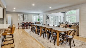 Ein geräumiger Essbereich mit einem langen Holztisch und mehreren Stühlen, ein Küchenbereich mit weißer Arbeitsplatte und Barhocker sowie eine gemütliche Sitzecke mit Sofa und Kissen.