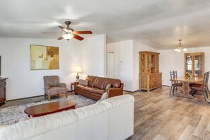 A living room with a brown leather sofa, a patterned armchair, a white sofa, a wooden coffee table, a dining table with chairs, a wooden cabinet, and a ceiling fan. - Magnolia Manor House: Desert Escape, Mtn Views (Dolan Springs)