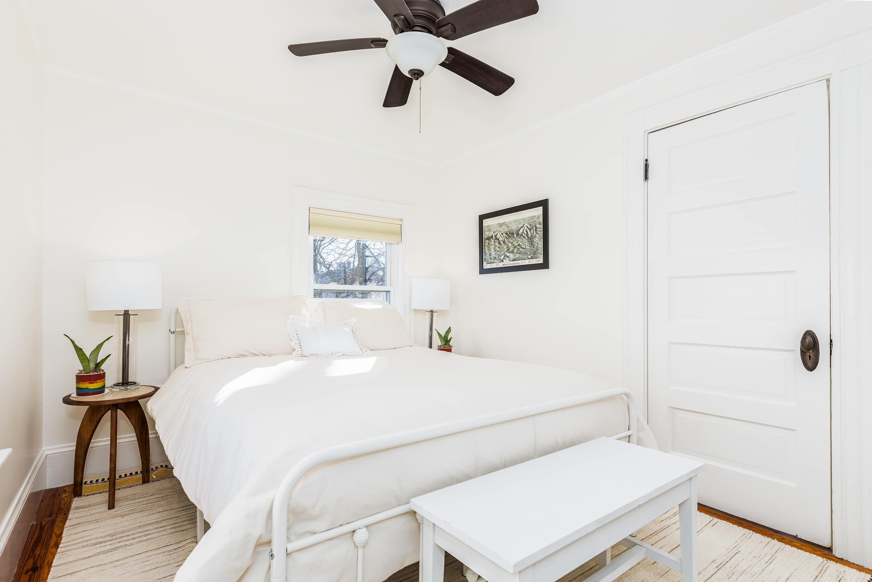Quarto com cama, mesas de cabeceira, ventoinha no teto, janela e porta.