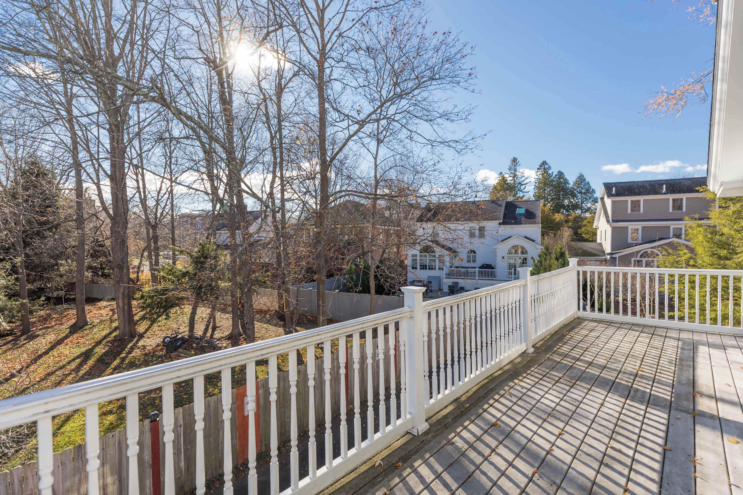 Une terrasse en bois avec une balustrade blanche, donnant sur un quartier résidentiel avec des maisons et des arbres couverts de feuilles.