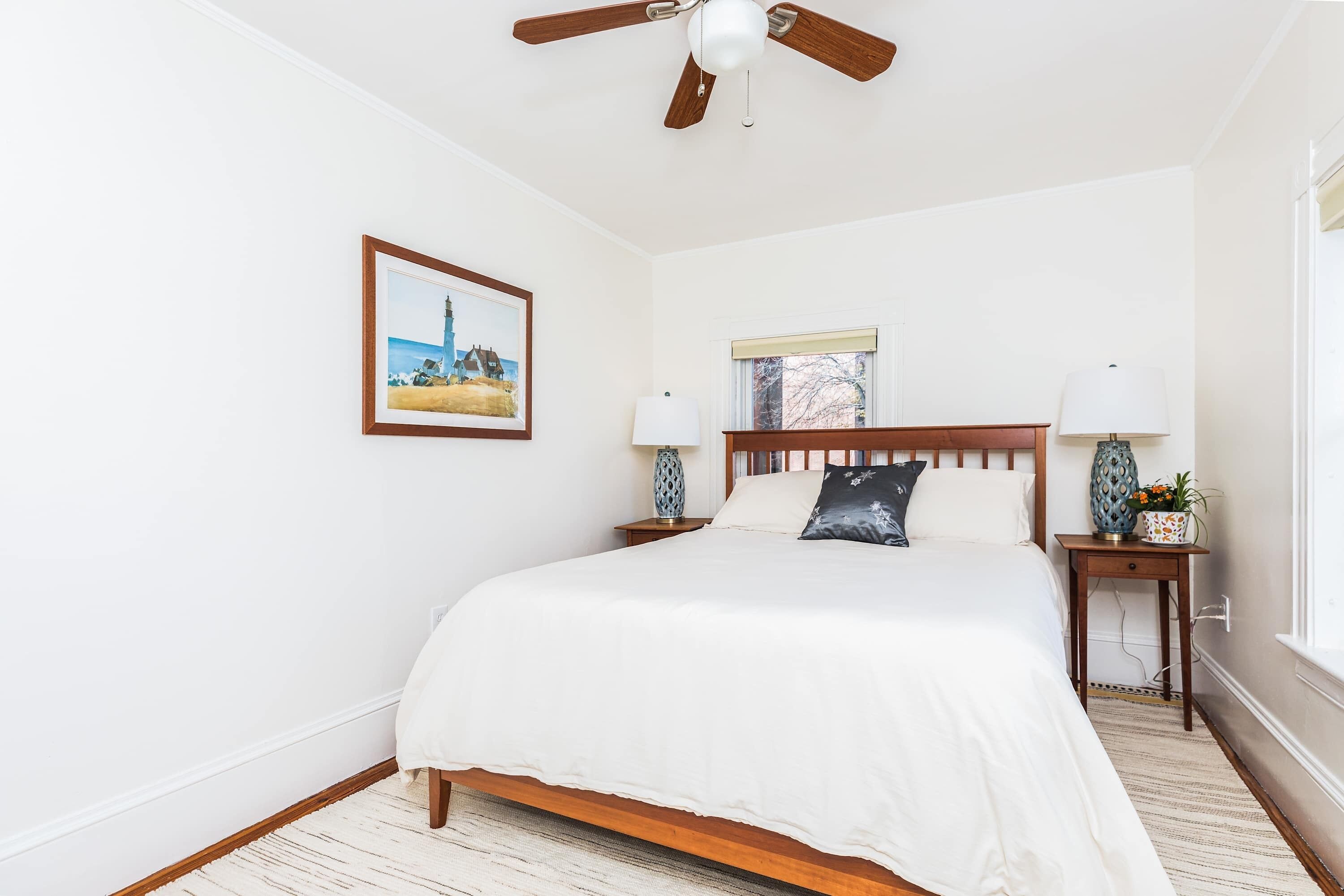 A bedroom with a wooden bed, white bedding, a ceiling fan, a nightstand with a lamp, a framed picture on the wall, and a window with a view of trees.
